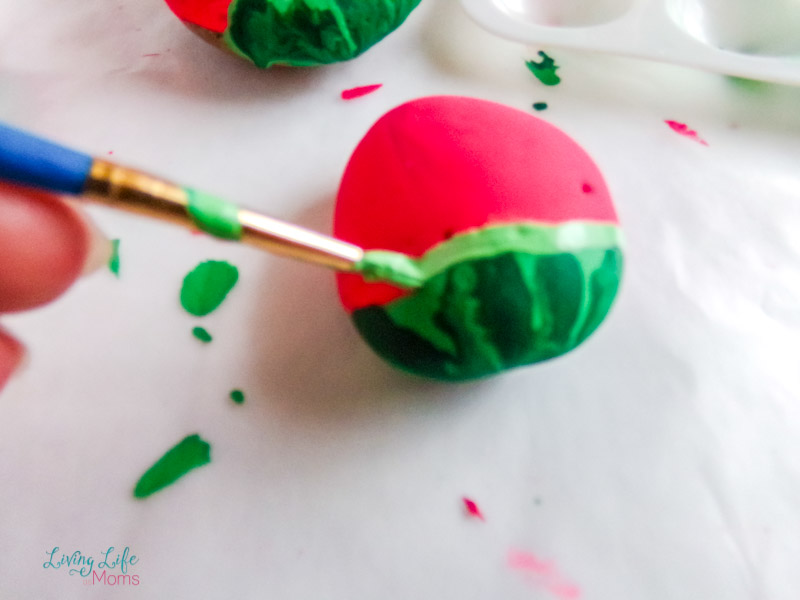 Adding light green paint to the border of the watermelon painted rock between the red and green paint after it has dried